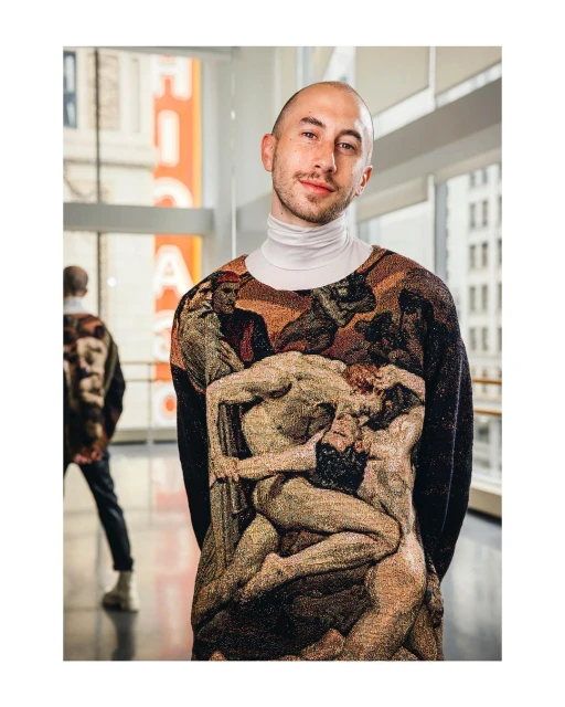 Portrait of a young adult man in a unique sweater with artwork, captured indoors with natural light and a blurred background figure.