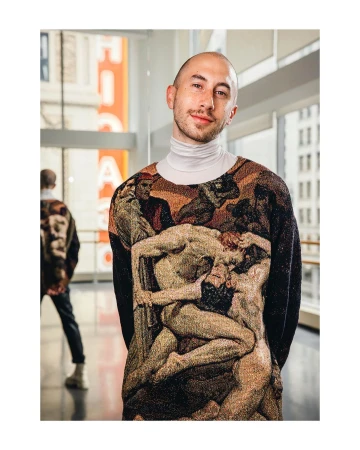Portrait of a young adult man in a unique sweater with artwork, captured indoors with natural light and a blurred background figure.