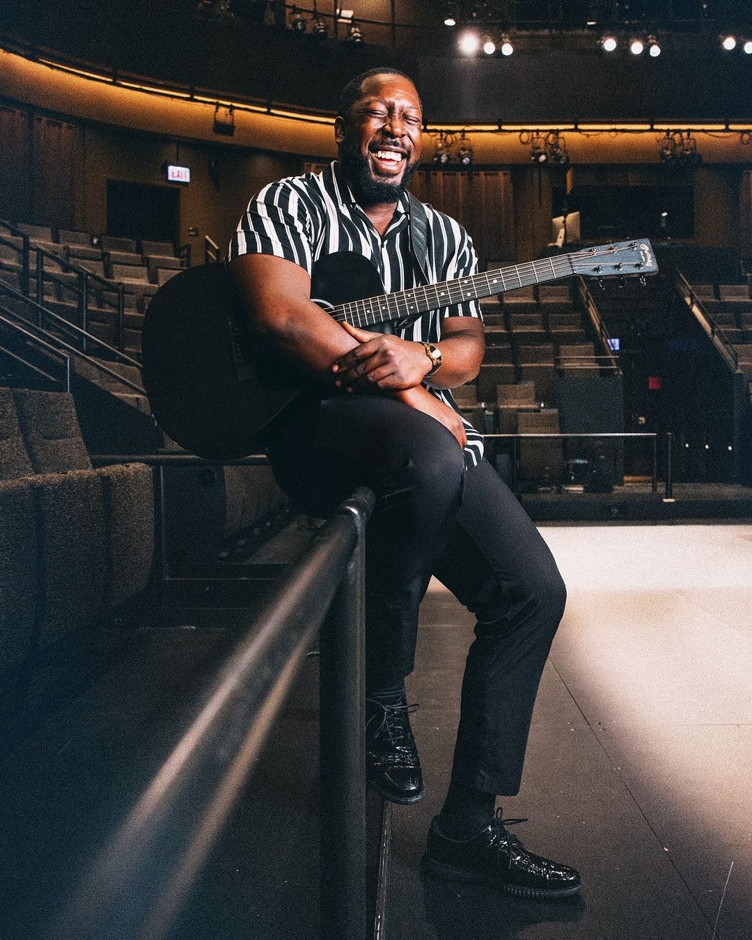 Smiling African-American man sitting on a railing, playing an acoustic guitar in an empty theater with moody, low lighting.