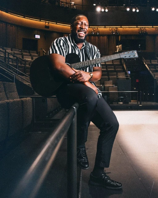 Smiling African-American man sitting on a railing, playing an acoustic guitar in an empty theater with moody, low lighting.