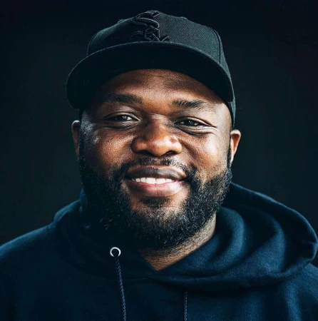 Headshot of an adult male with a dark complexion wearing a baseball cap and a hooded sweatshirt against a dark background.