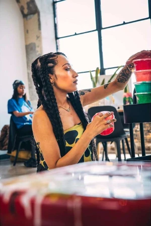 Woman with braided dark hair wearing a patterned top holding a small pink container inside a bright studio space