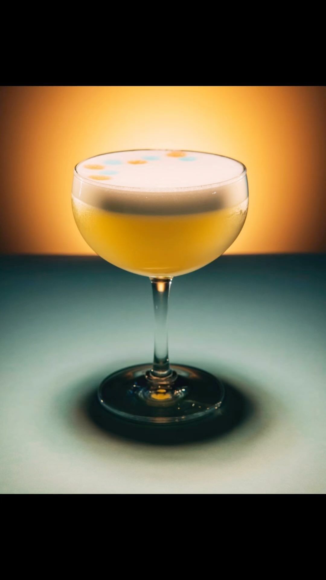 A stemmed cocktail glass filled with a yellow drink and a white foamy top is illuminated against a gradient yellow and blue backdrop.
