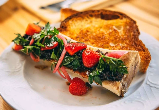 Plated bone marrow with herbs, strawberries, and toast, showcasing culinary artistry and a rich, savoury-sweet flavor combination.