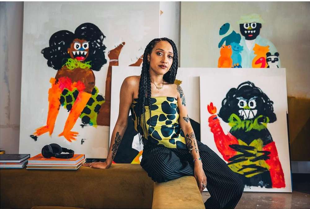 Artist in studio poses with vivid paintings. Woman with braids sits near colorful abstract art on canvas and books, wearing a printed top.
