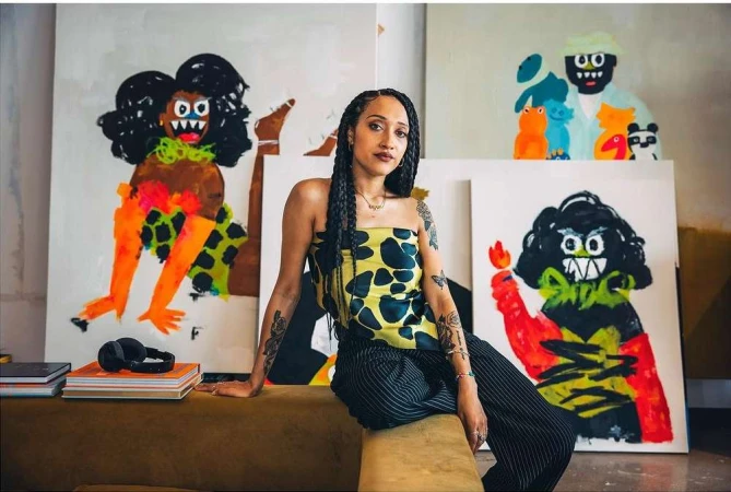 Artist in studio poses with vivid paintings. Woman with braids sits near colorful abstract art on canvas and books, wearing a printed top.