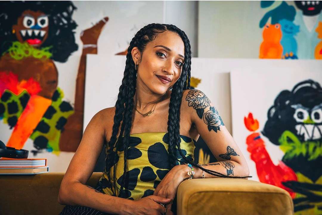 Portrait of a woman with braided hair sitting in front of paintings in an art studio wearing a patterned dress with tattoo on arm.