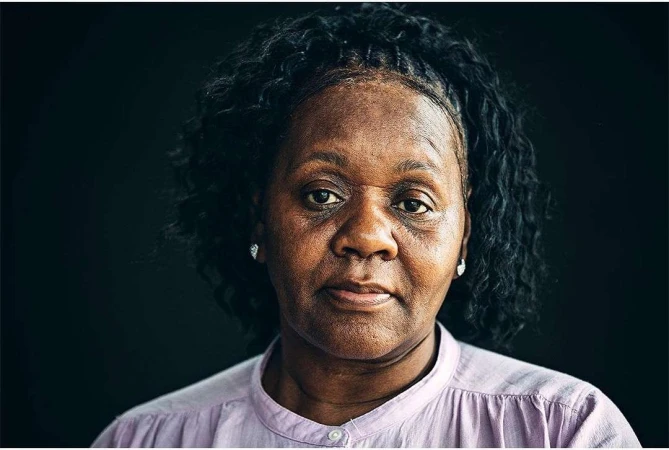 Close-up portrait of a Black woman with dark curly hair wearing a light pink shirt against a dark background
