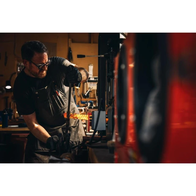 A skilled adult male artisan in safety glasses works with molten glass at a metal machine in a workshop with natural lighting.