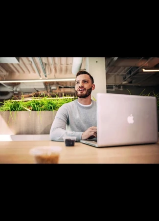 Man seated at a wooden desk with a silver laptop in an open plan office space