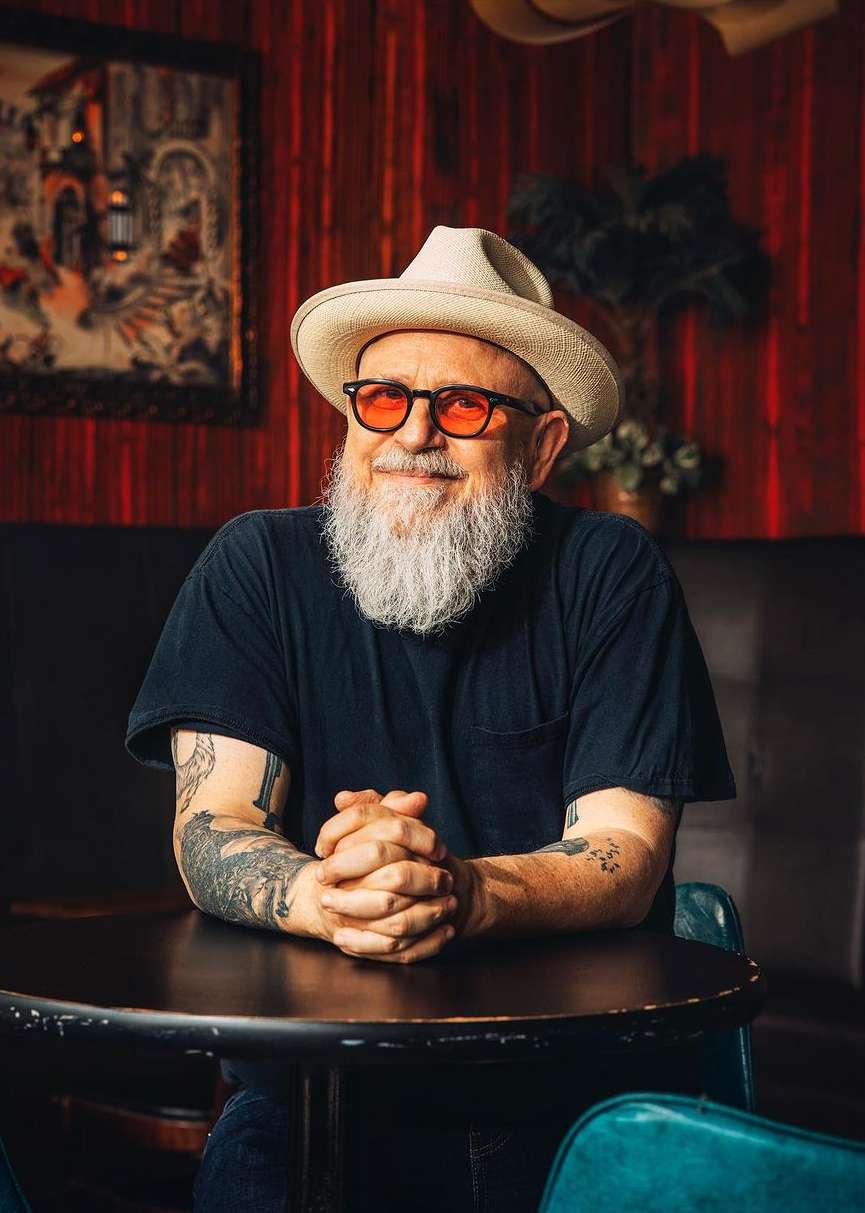 An older adult man with a white beard and a hat poses at a dark table indoors, wearing orange glasses and a dark T-shirt.
