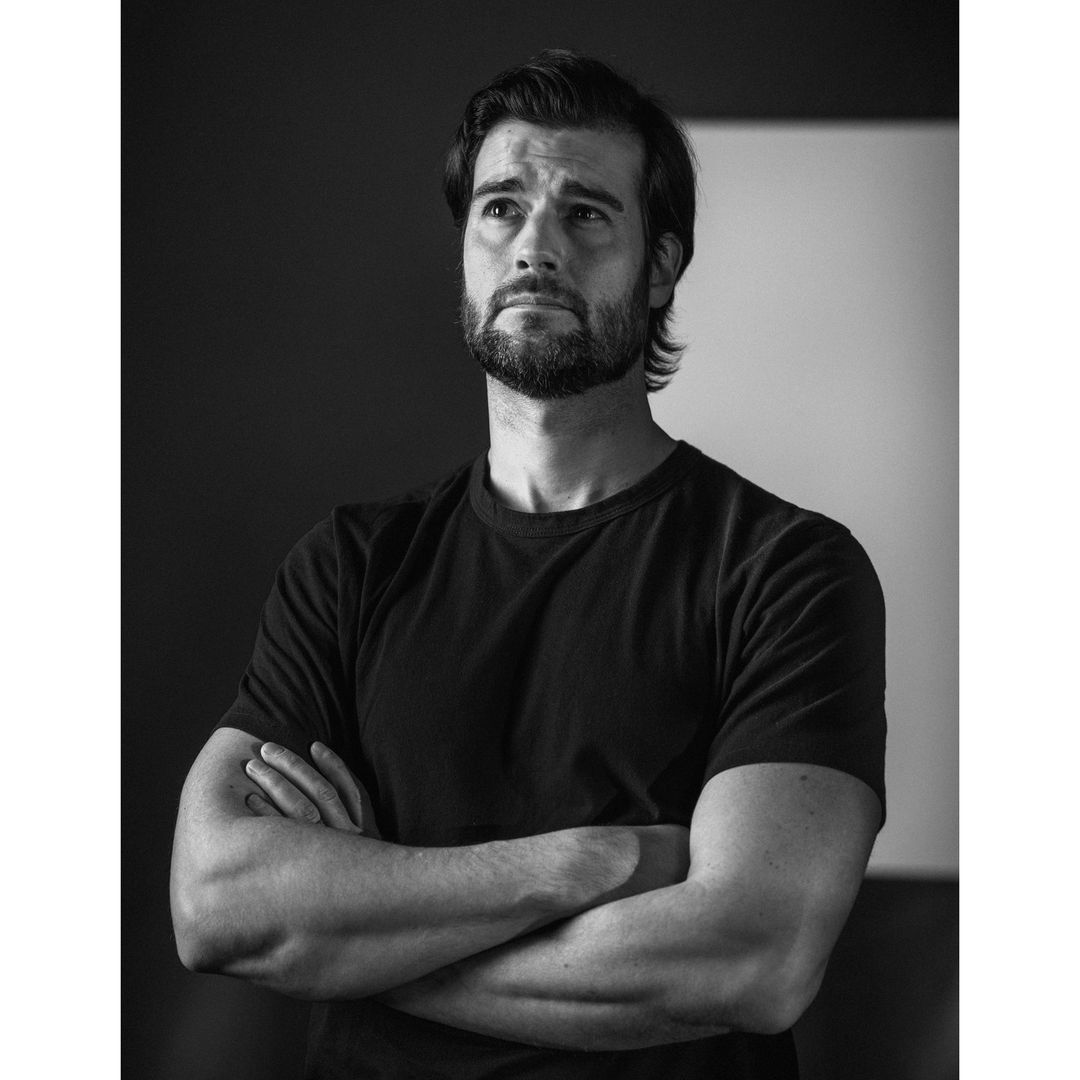 Monochrome portrait of a bearded man in a black T-shirt with crossed arms, looking up against a dark and light grey background.