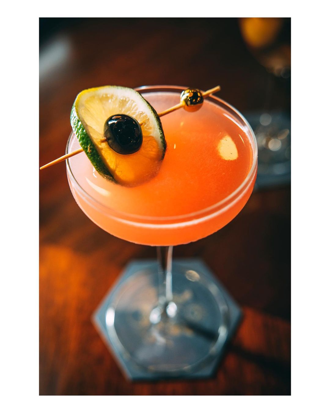 Close-up of a vibrant pink cocktail in a stemmed glass, garnished with a lime slice and cherry, set on a dark wooden table.