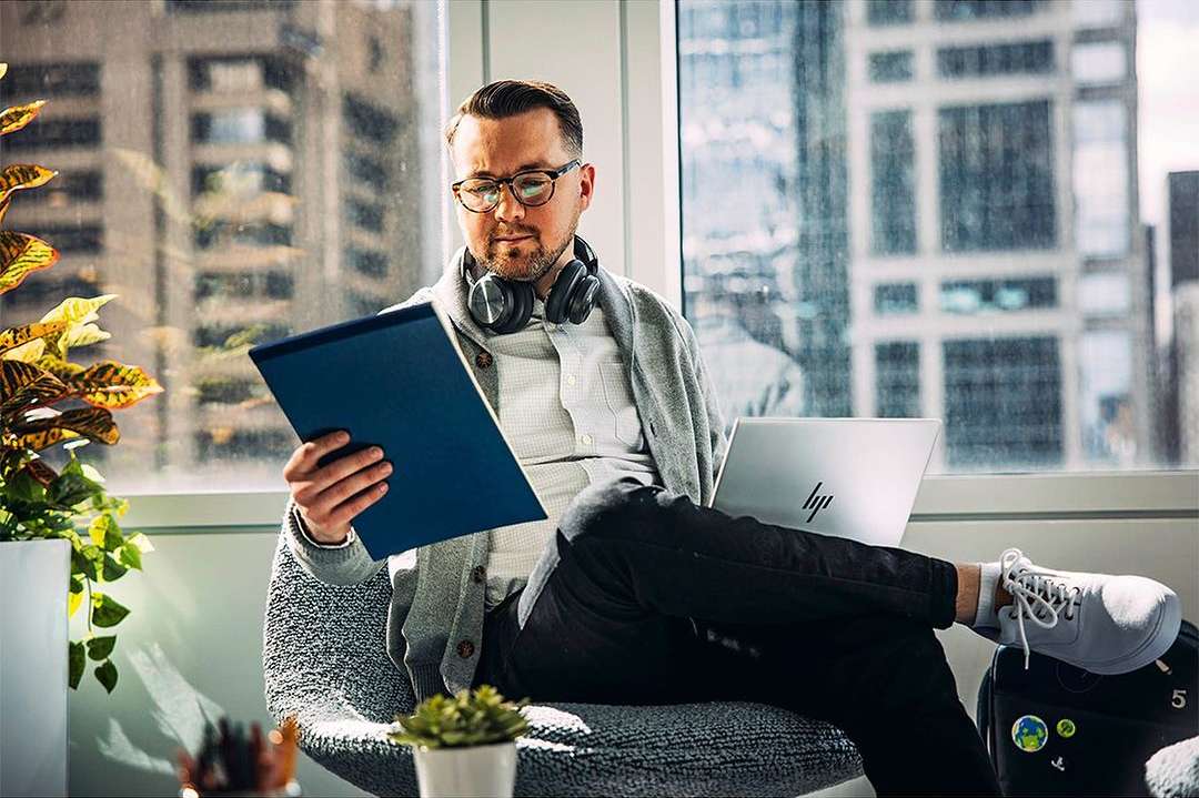 Man with glasses and headphones sits in modern office, reading a blue tablet with a laptop, plants, and a cityscape view.