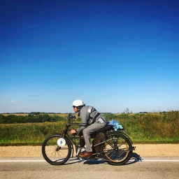 Man wearing a white helmet and gray riding gear riding a vintage motorcycle along a road against a clear blue sky