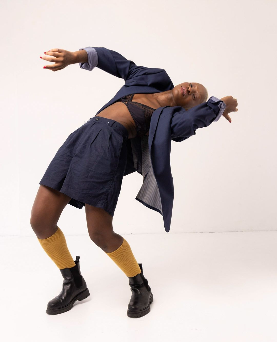 A bald Black model with a lace bra, oversized blue blazer and shorts, and gold socks poses dynamically against a white background.