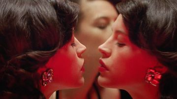 Close-up of two women with dark curly hair and red lighting, wearing earrings. One woman slightly blurred in the background.