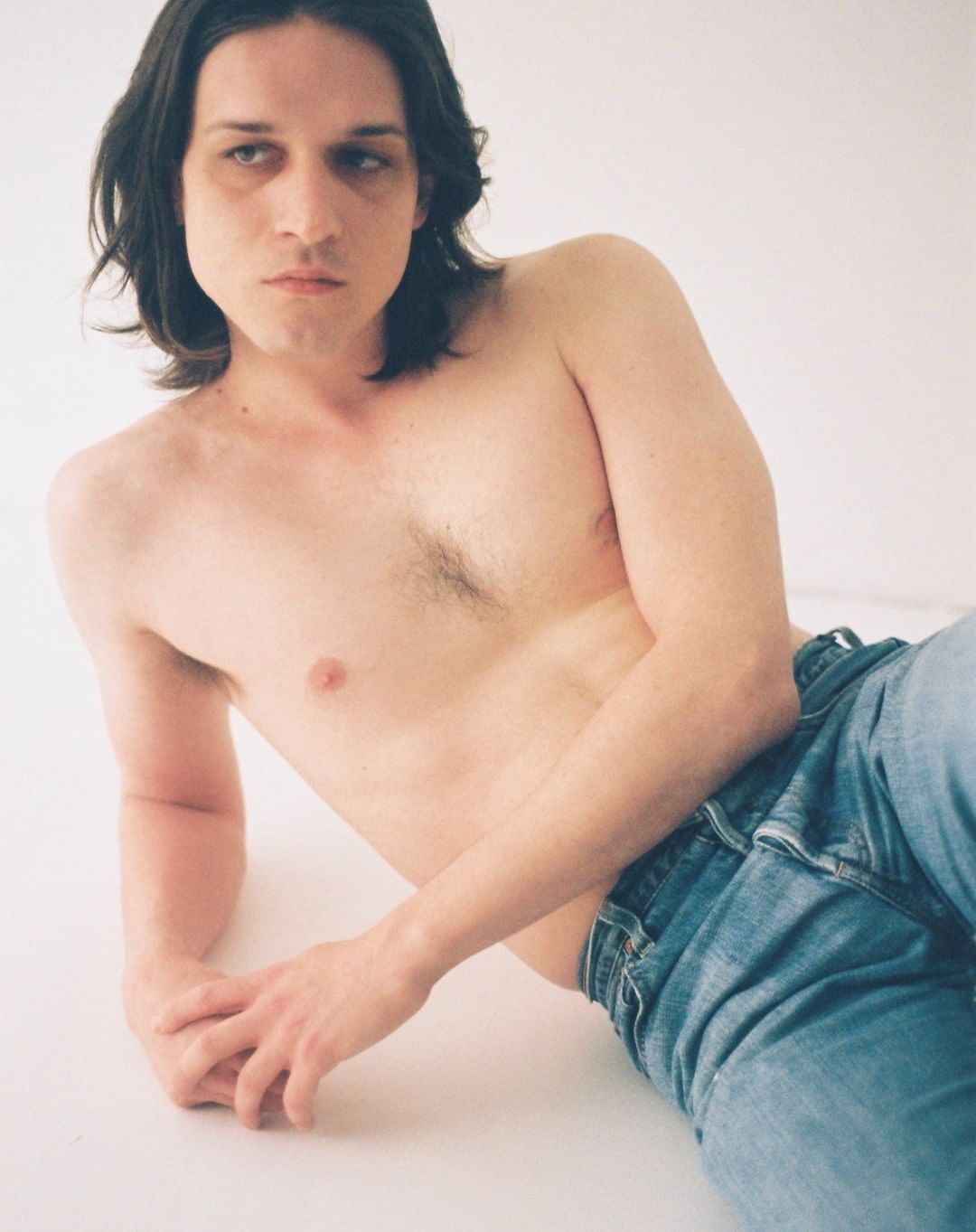 Shirtless man with long dark hair reclines in jeans against a white background, gazing directly at the camera in a studio shot.