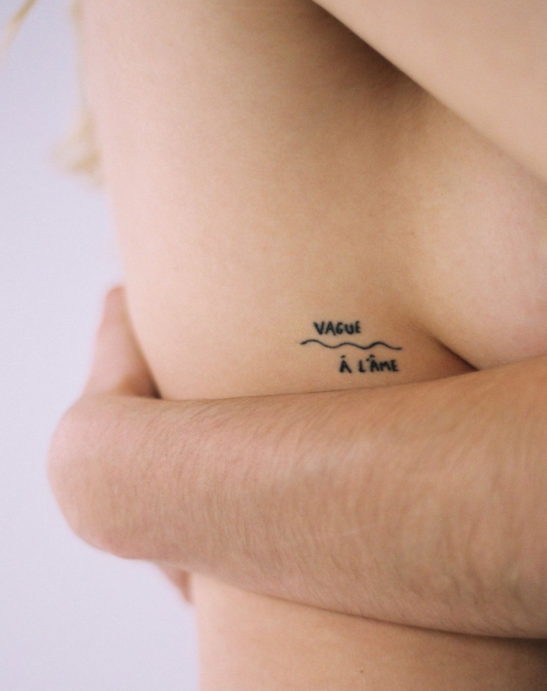 Close-up studio shot featuring a person's torso with a tattoo, their arms gently folded across their body, soft studio lighting.