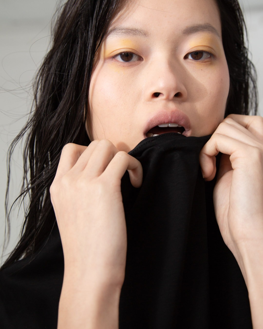 Close-up portrait of an Asian woman with dark hair pulling on a black shirt and looking at the camera with light makeup.