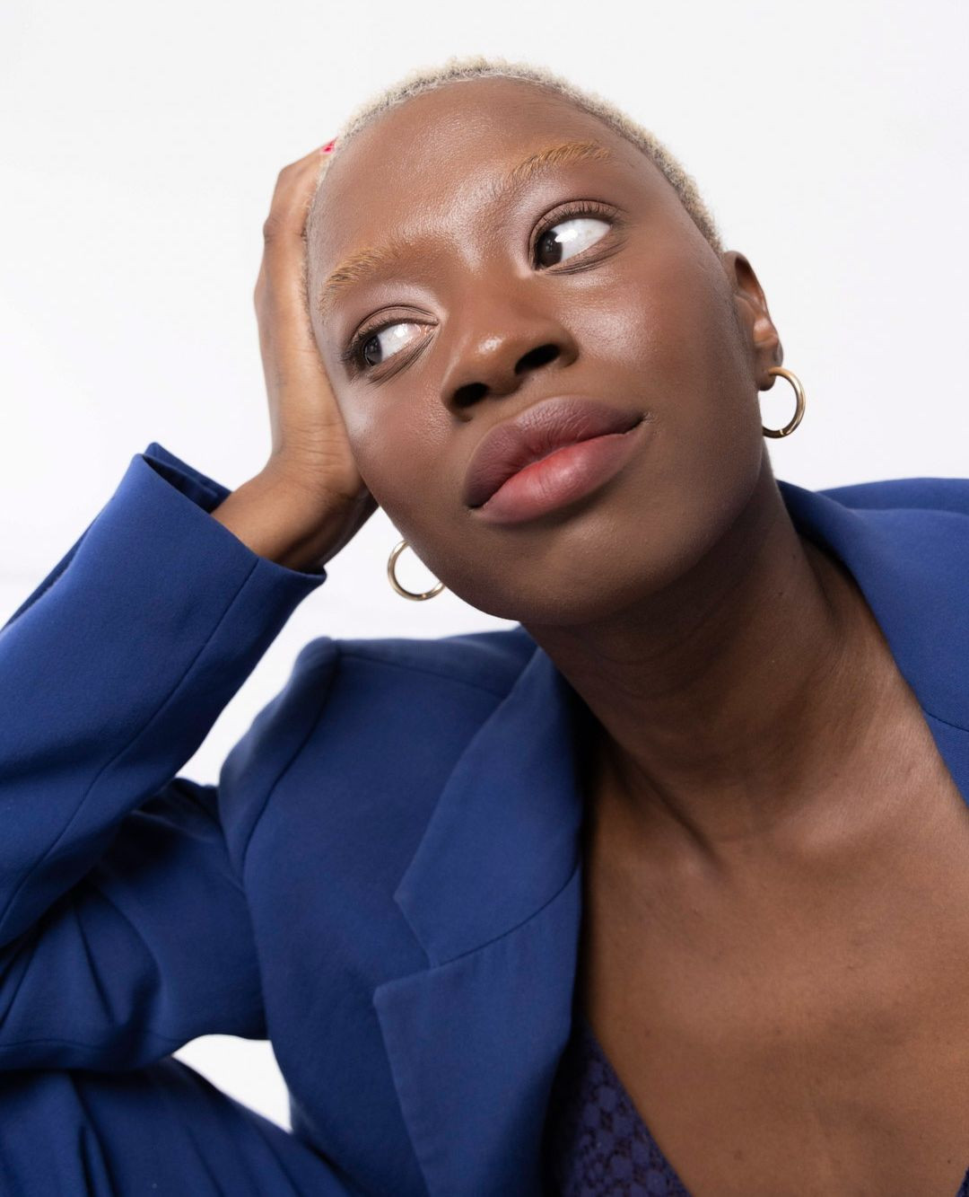 Portrait of a young African woman with a close-up view. She has short blonde hair, wears gold hoop earrings, and a blue blazer against a white background.