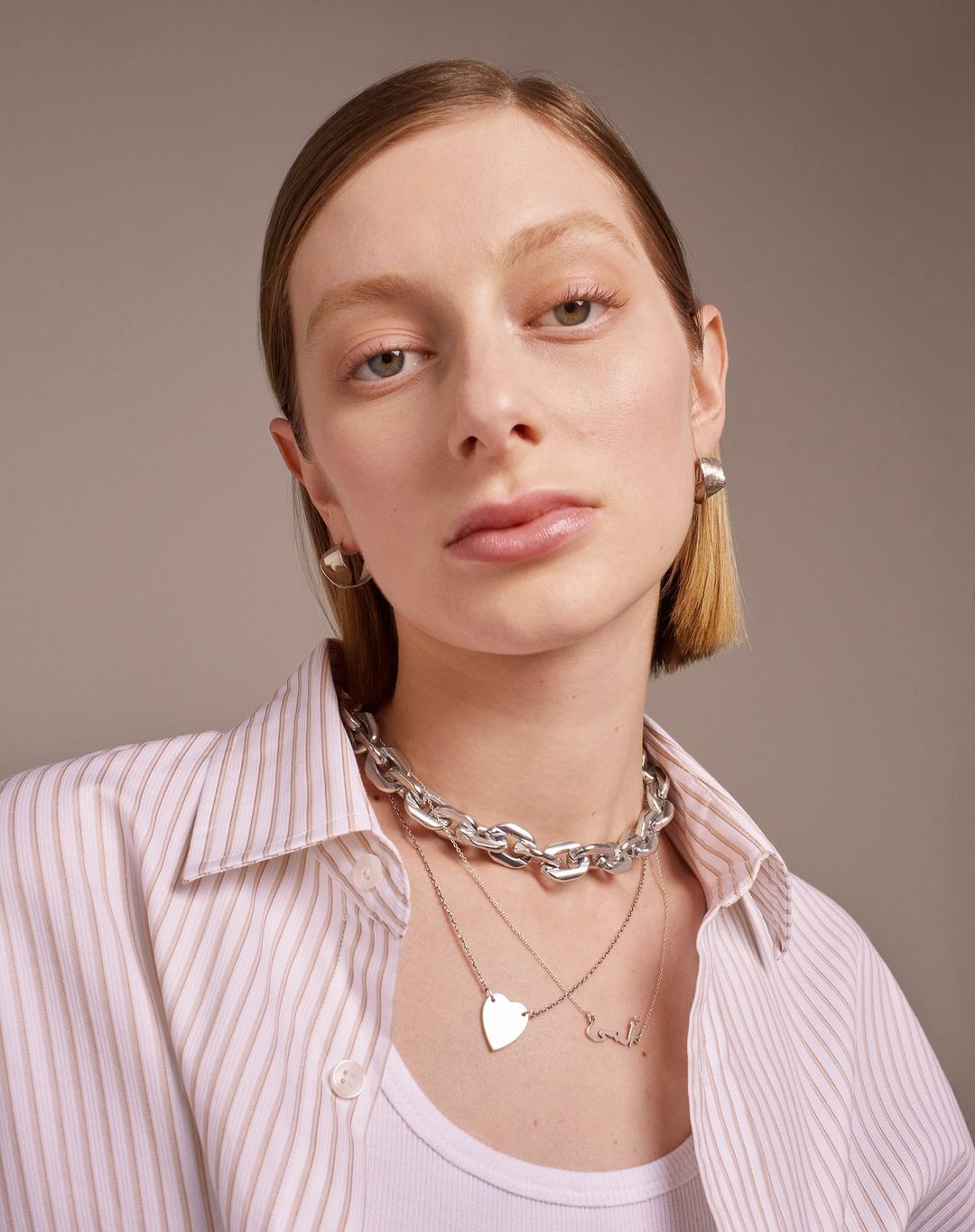 Portrait of a young woman with short blonde hair wearing a button-down shirt, silver jewelry, and light makeup against a neutral background.