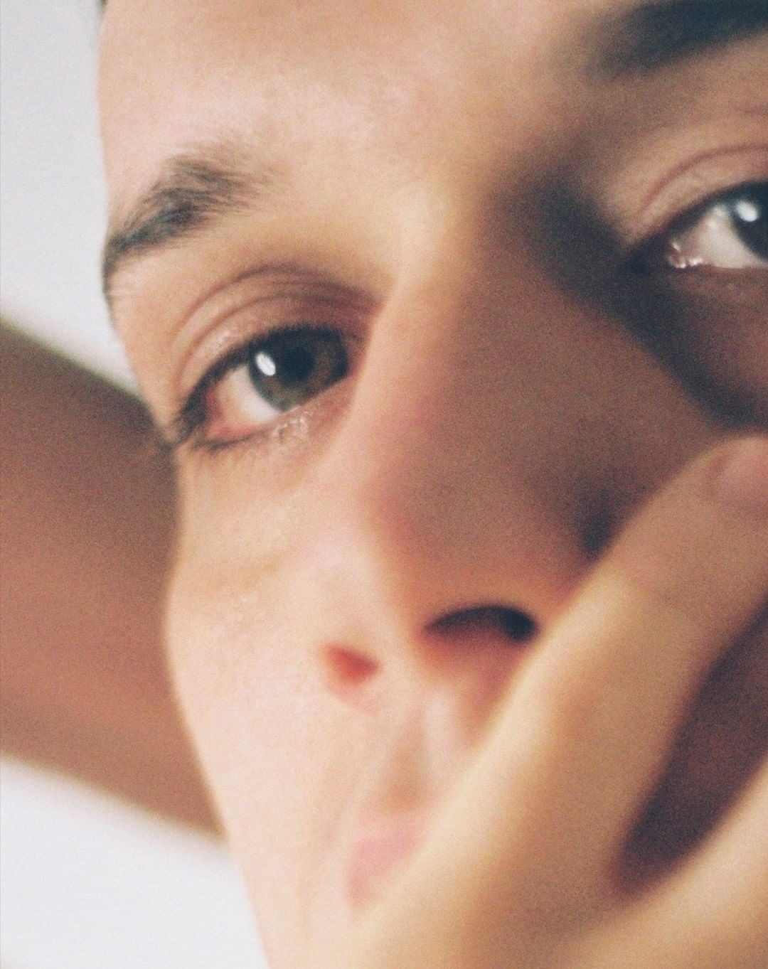 Close-up portrait of a young adult man with his hand near his face, showcasing natural light and shallow depth of field on a plain background.