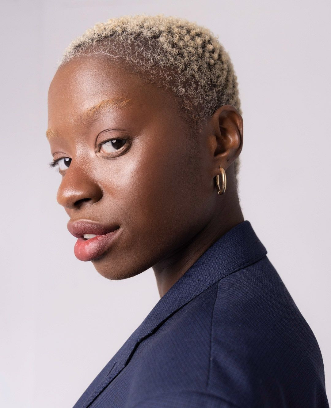 Close-up portrait of a young adult woman with short bleached blonde hair, wearing a navy blazer, in a studio setting with soft lighting.