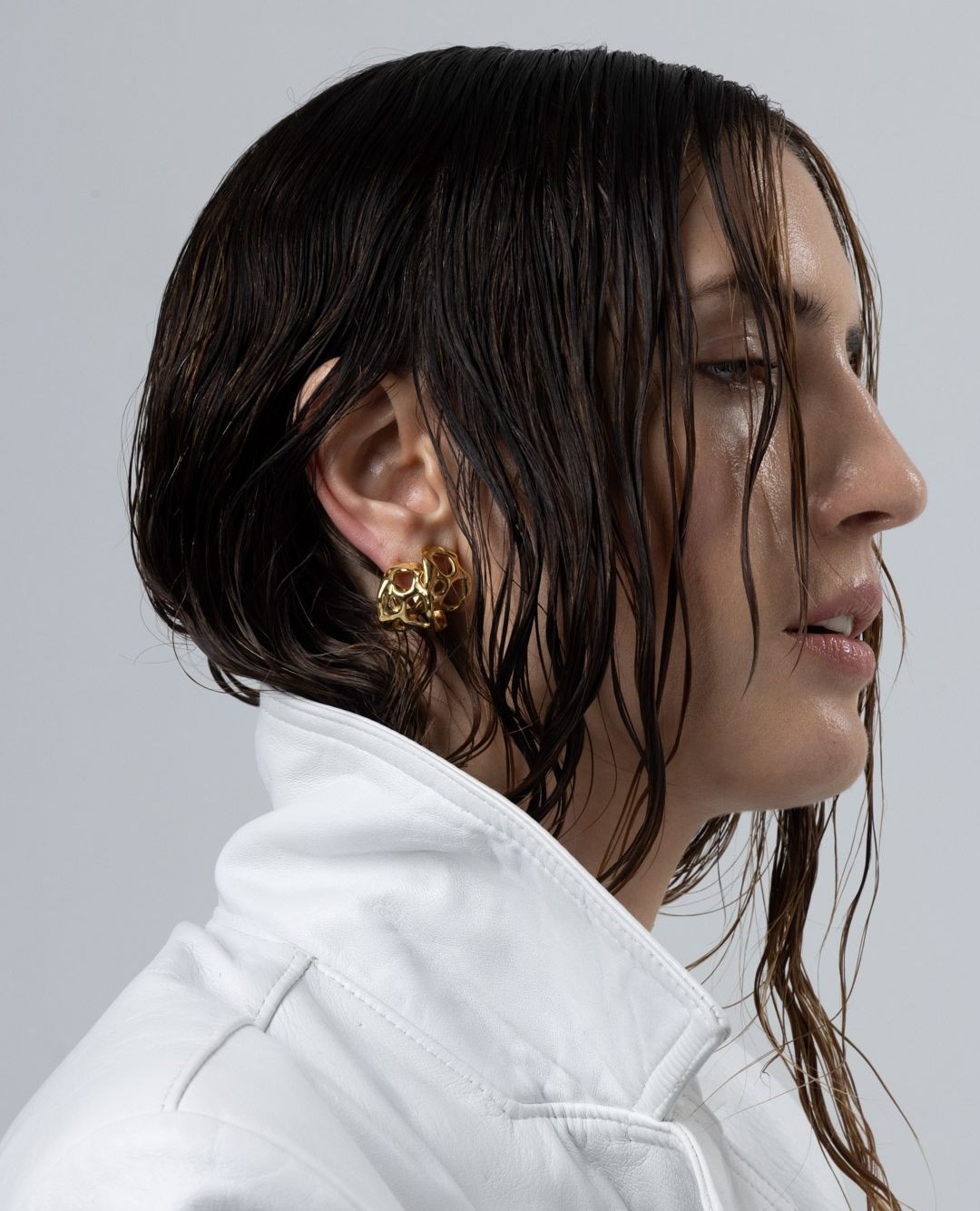 Profile view of an adult woman with slicked-back dark hair, wearing a white jacket and gold earrings against a plain gray backdrop.