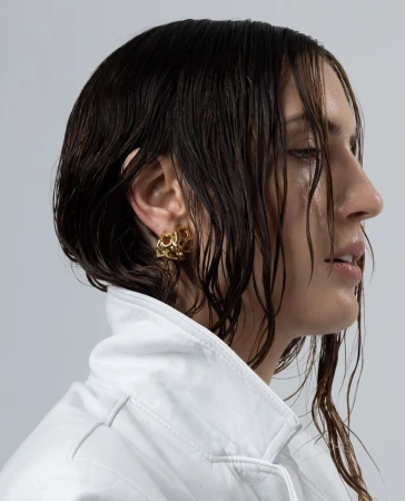 Close-up profile portrait of a woman with wet dark hair wearing a white high collar jacket and gold textured earrings.