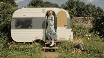 A woman exits a vintage camper in a rural setting on a sunny day, wearing a long floral dress and white boots. The scene evokes a vintage travel aesthetic.