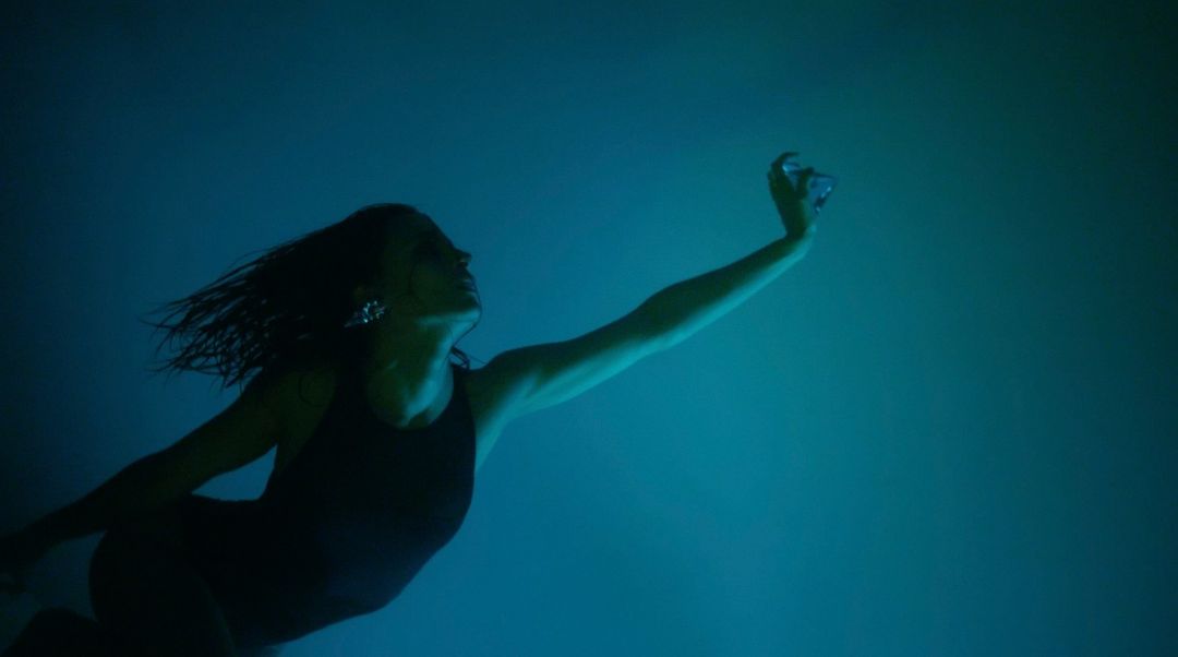 Woman underwater wearing a dark swimsuit with her arm raised up, captured in deep blue and teal tones by photographer Clemence Demesme.