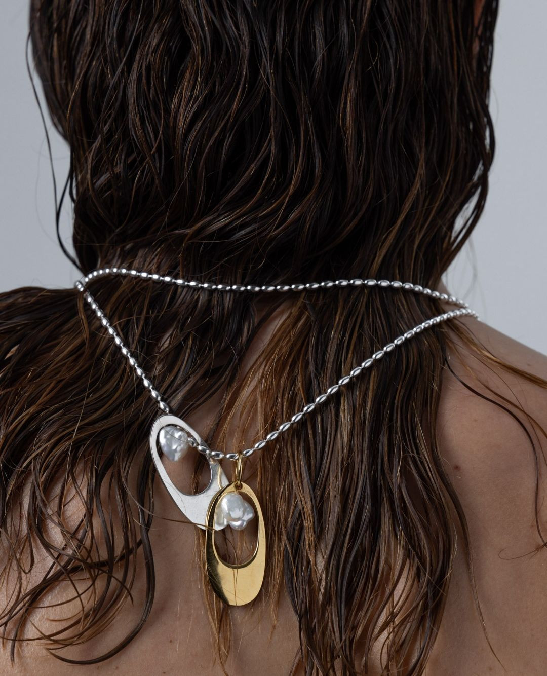 Close-up studio shot of a woman with wet brown hair wearing a silver beaded necklace with white and gold pendant charms.