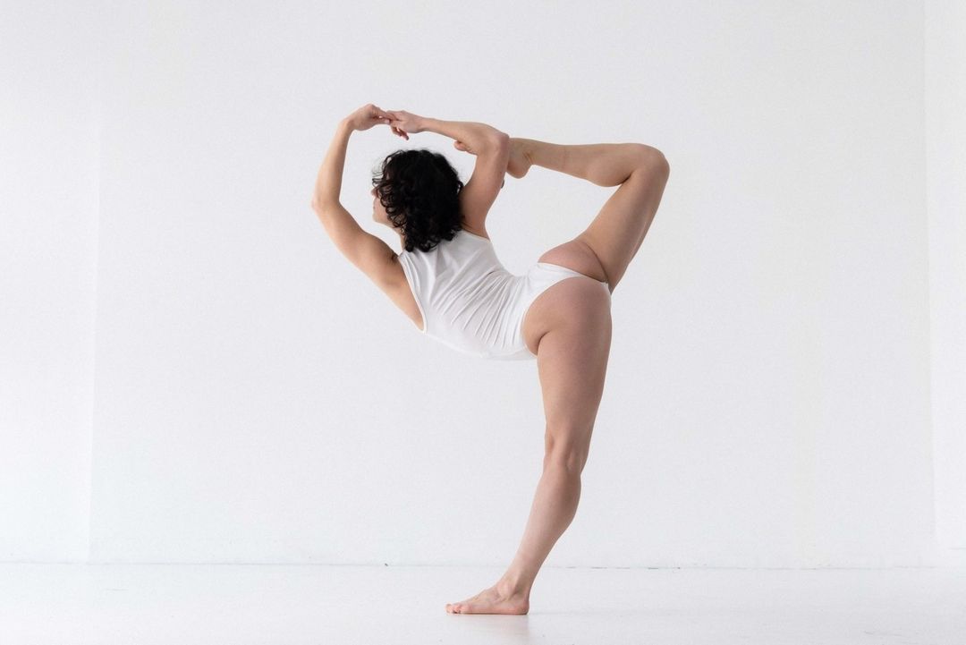 Woman in white bodysuit doing yoga pose against a white wall. Fitness and wellness concept showcasing strength and flexibility.