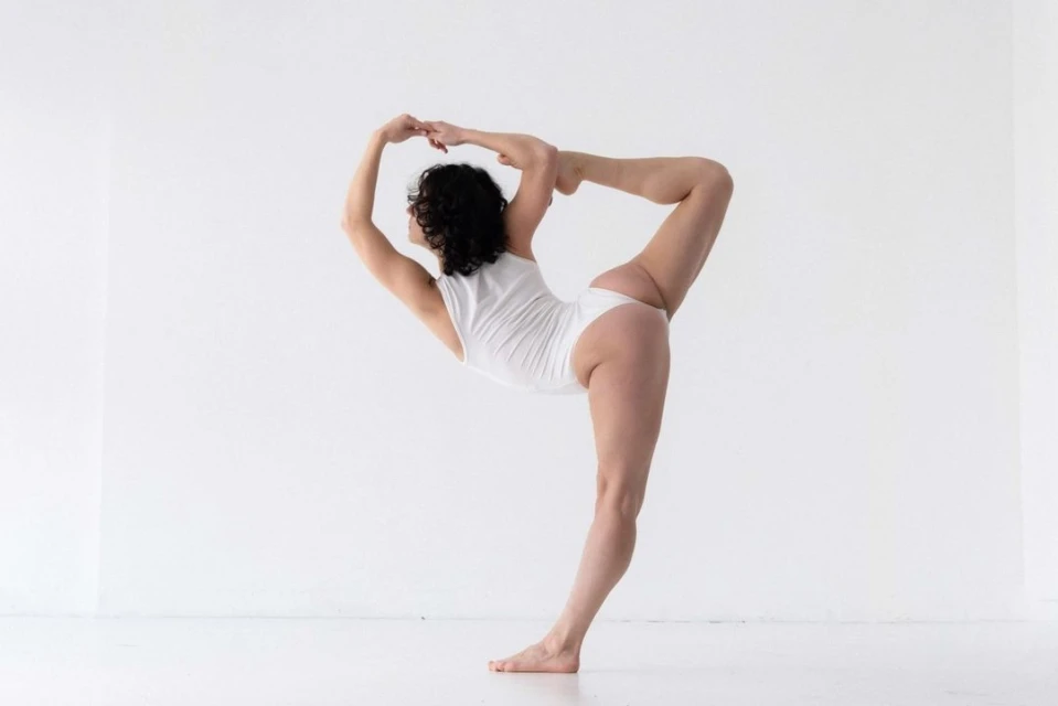 Woman in white bodysuit doing yoga pose against a white wall. Fitness and wellness concept showcasing strength and flexibility.