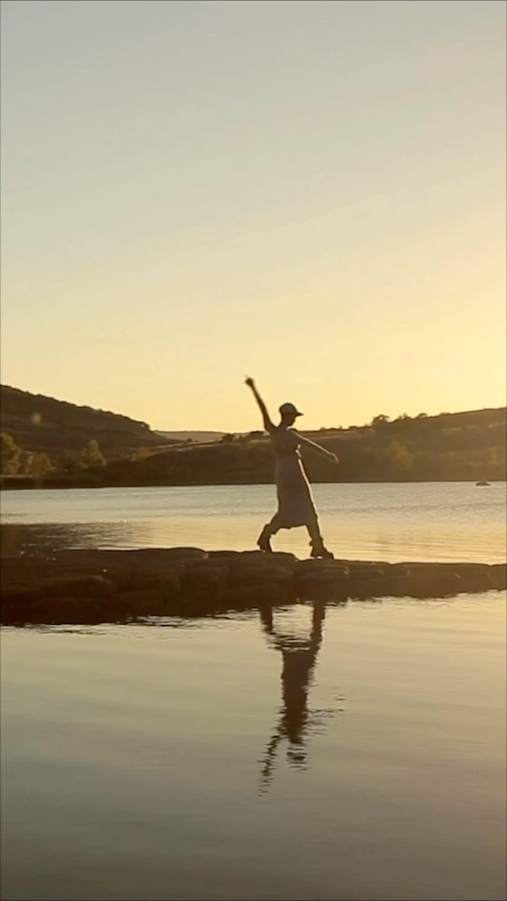 Person in dress and hat balancing on a stone breakwater with a lake reflecting the figure at golden hour with hillside in the background.