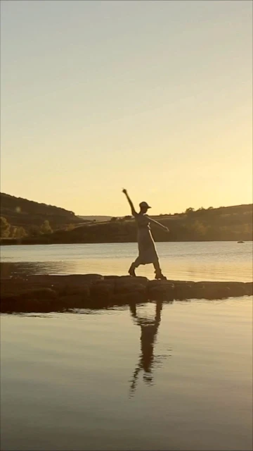Person in dress and hat balancing on a stone breakwater with a lake reflecting the figure at golden hour with hillside in the background.