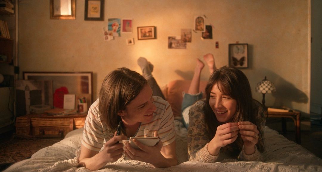 Two smiling adults, a male and a female, are lying on a bed in a warmly lit room with photos on the wall and a homey atmosphere.