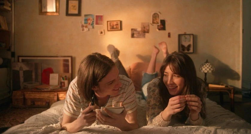 Two smiling adults, a male and a female, are lying on a bed in a warmly lit room with photos on the wall and a homey atmosphere.