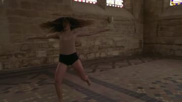 A slender woman with flowing hair is dancing in a classical interior, wearing a nude top and dark bottoms, lit by daylight.