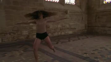 A slender woman with flowing hair is dancing in a classical interior, wearing a nude top and dark bottoms, lit by daylight.