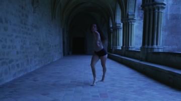 A barefoot young adult female in active pose in an arched stone hallway, bathed in blue light in a historic building's architecture.
