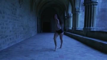 A barefoot young adult female in active pose in an arched stone hallway, bathed in blue light in a historic building's architecture.