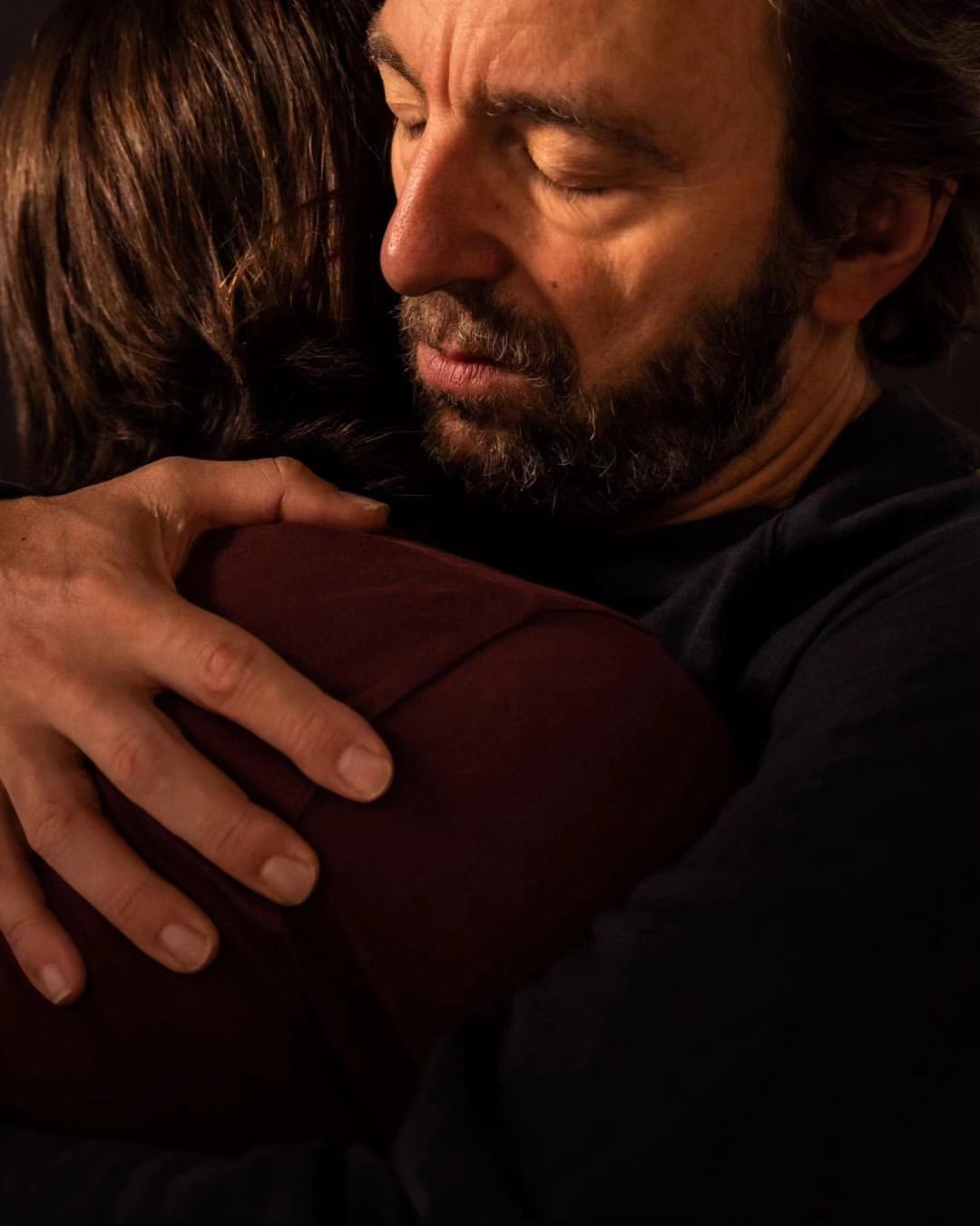 Close-up of a man embracing another person, with his eyes closed and a gentle expression, conveying comfort and warmth in a dark indoor setting.