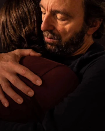 Close-up of a man embracing another person, with his eyes closed and a gentle expression, conveying comfort and warmth in a dark indoor setting.