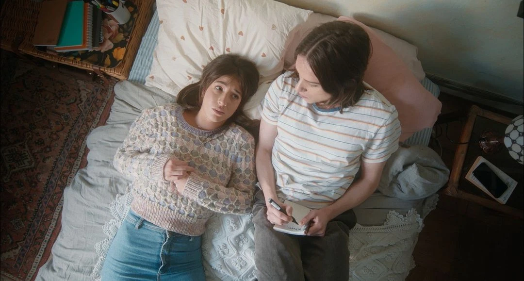 Two adults, a woman and a man, lie side-by-side on a patterned bed; the man is writing in a notebook in a cozy, indoor setting.