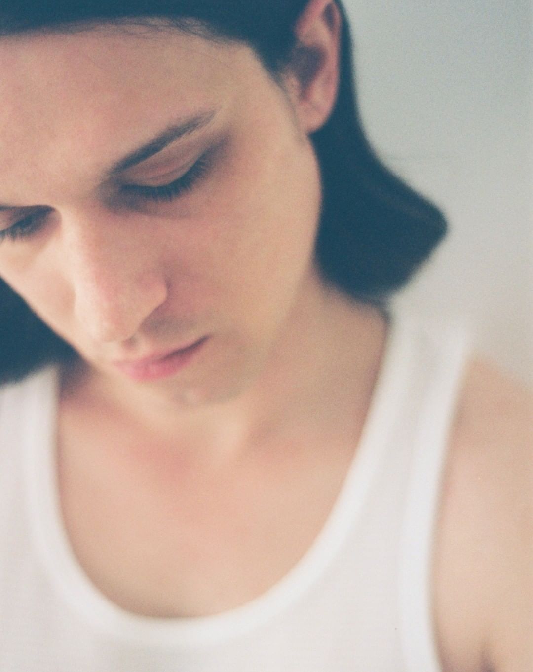 Close-up portrait of a young adult with long dark hair, wearing a white tank top, looking down in an indoor setting with soft lighting.