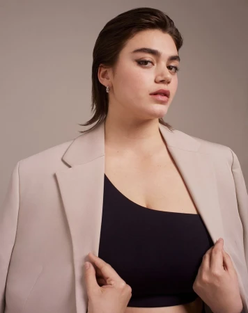 Portrait of a woman with slicked-back hair wearing a beige blazer over a black top against a solid neutral background.