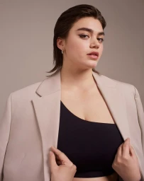 Portrait of a woman with slicked-back hair wearing a beige blazer over a black top against a solid neutral background.