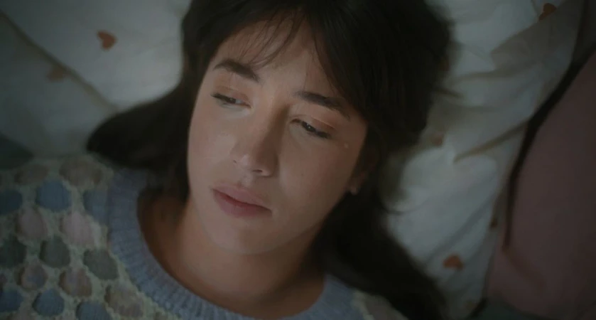 Close-up portrait of a woman with dark hair lying on patterned pillows wearing a textured knit sweater by Clemence Demesme.